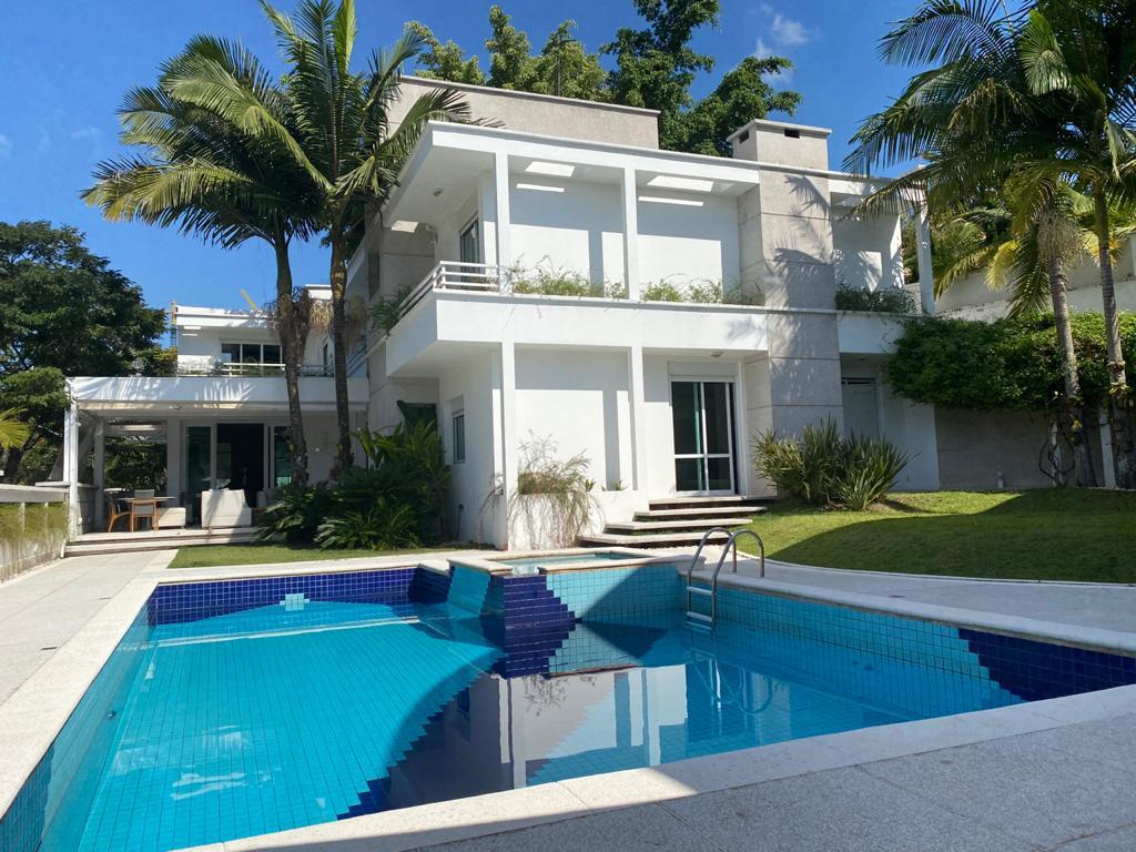 A modern two-story house is shown, featuring large glass windows and a balcony. A swimming pool with a blue tiled surface is located in the foreground, surrounded by palm trees and lush greenery, creating a serene outdoor environment.