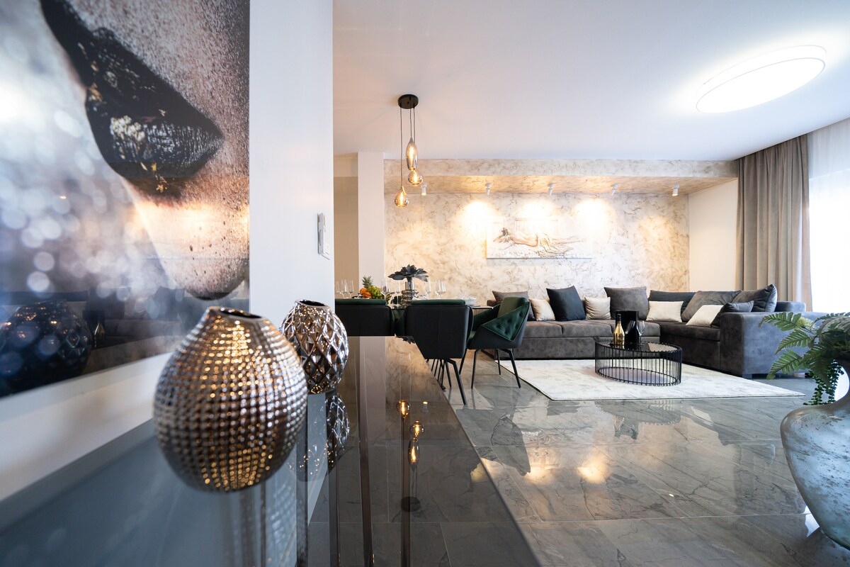 A modern living area features a large sectional sofa, complemented by a round coffee table and elegant lighting. Decorative accents, including metallic vases, enhance the space. A textured artwork adds visual interest, while floor-to-ceiling windows allow natural light to fill the room.
