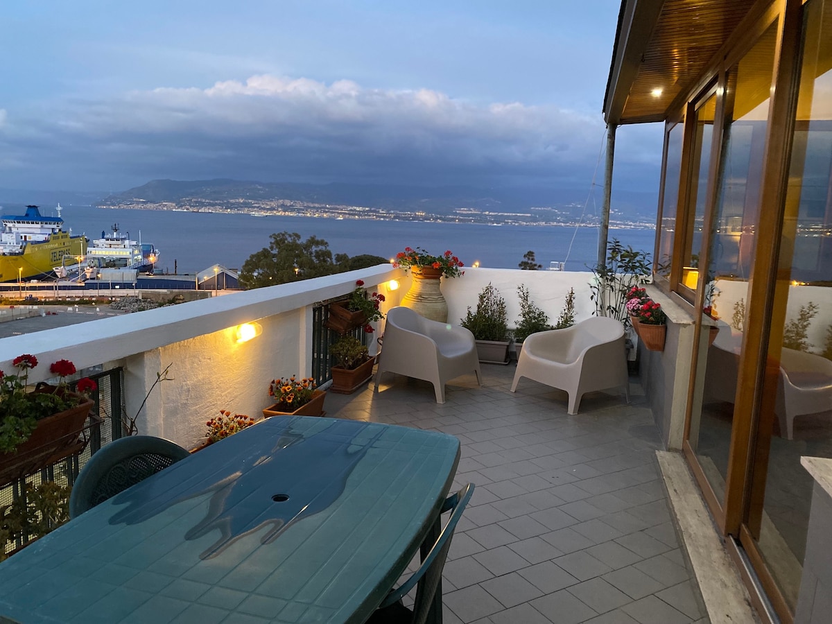 An inviting terrace offers views of the Messina Strait, surrounded by various potted plants. Two white chairs and a table create a cozy seating area, illuminated by soft lighting, ideal for enjoying evening moments or morning coffee.