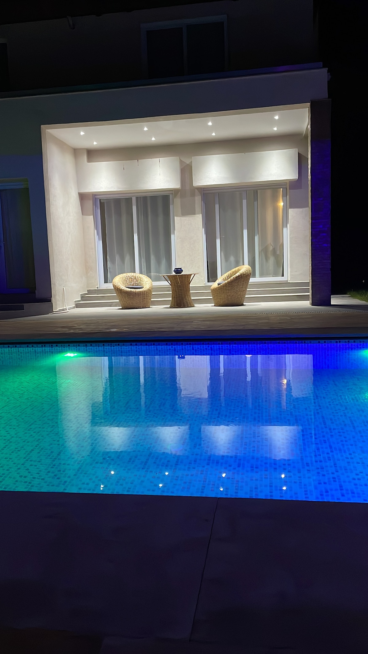 An illuminated pool area features two wicker chairs positioned by the water's edge. The serene space is enhanced by soft lighting, reflecting off the pool's surface. Sliding glass doors lead to an indoor seating area, creating a blend of indoor and outdoor living.