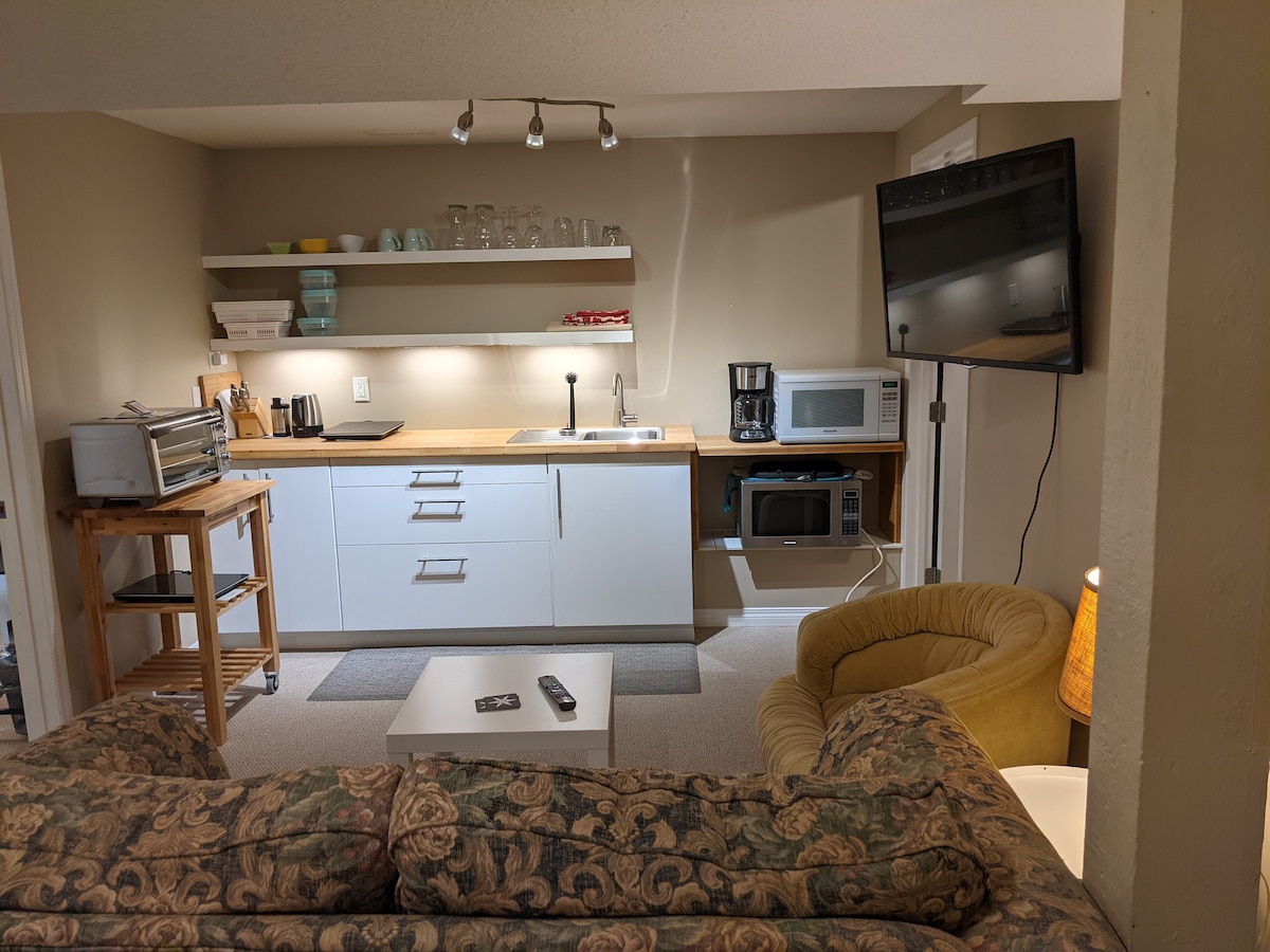 The kitchen area is equipped with modern cabinetry and a variety of kitchenware, including glasses and utensils. A coffee maker, microwave, and toaster are positioned on the countertop. The living area features a cozy couch, a coffee table, and a flat-screen TV mounted on a stand.