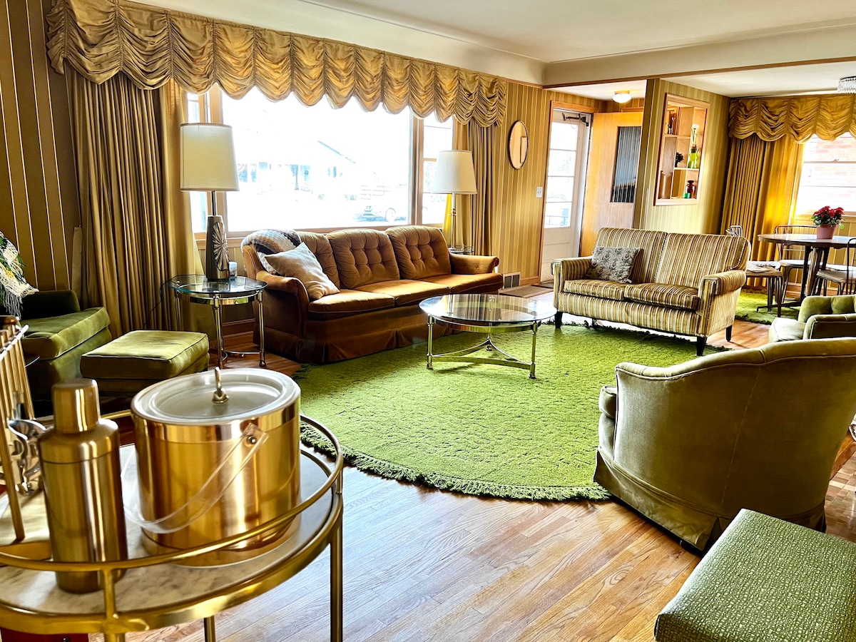 A spacious living area is furnished with mid-century sofas and armchairs, featuring warm gold and green tones. A glass coffee table sits on a large green area rug, while light streams through large windows dressed with gold drapes, creating a cozy atmosphere.