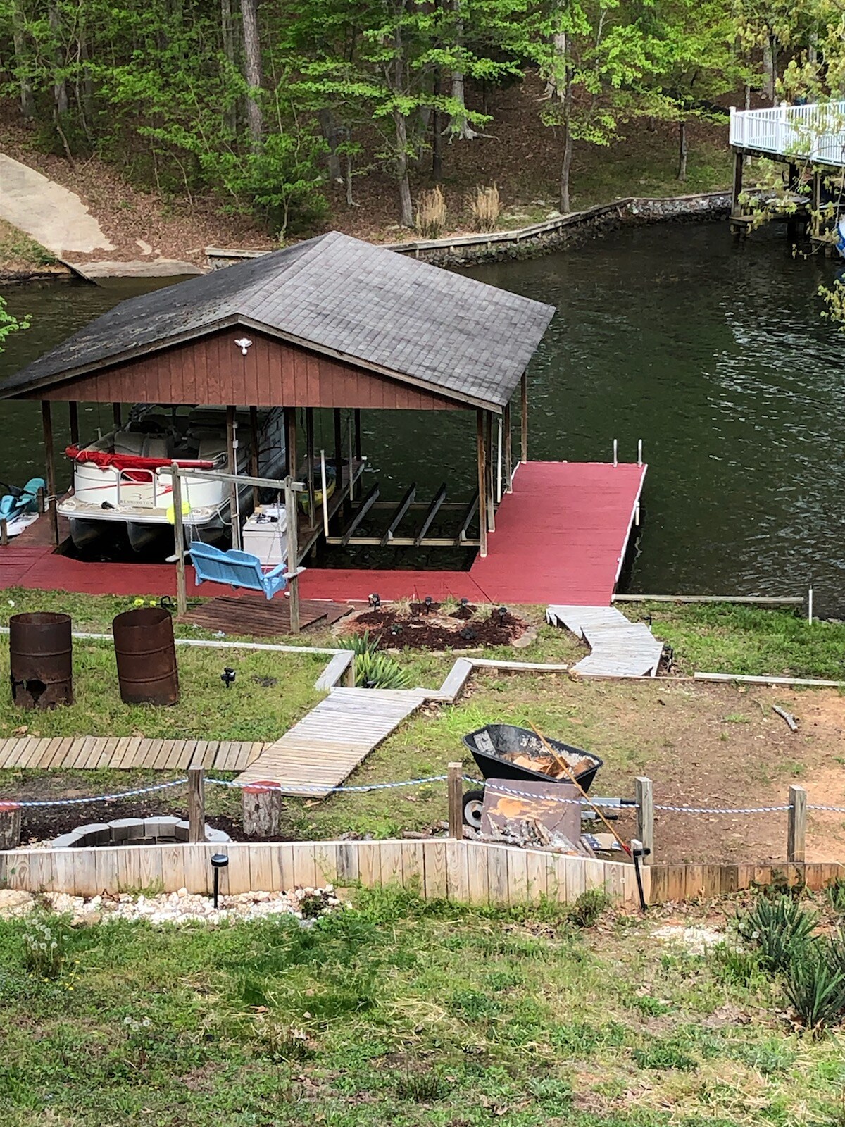 A boat house is positioned by the water, featuring a red deck and several covered spaces for boats. Adjacent pathways and a grassy area enhance accessibility. Two barrels are situated on the lower side, complemented by a fenced garden area with stones and lighting fixtures.