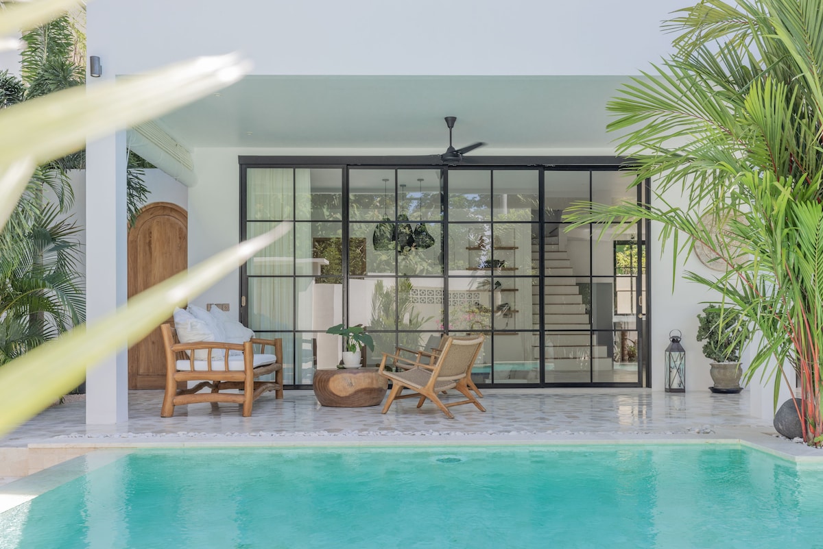 A modern villa features large glass doors opening to an indoor living space. Outdoor seating arrangements are positioned next to a private pool, while lush tropical plants frame the scene. Natural light filters in, highlighting the serene atmosphere of the area.