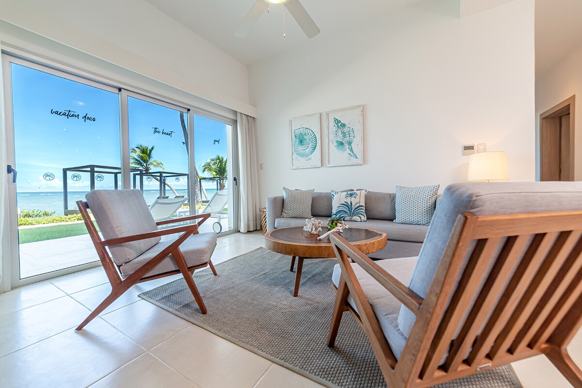 The living area is furnished with a comfortable gray sofa and two wooden chairs. A round coffee table is placed in the center, surrounded by a large area rug. Sliding glass doors provide a view of the beach and access to an outdoor space.
