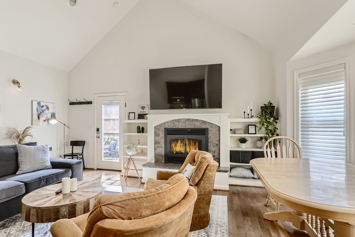 A spacious living area features a large gas fireplace and a mounted television. Comfortable seating includes a plush couch and two accent chairs. Natural light enters through nearby windows, illuminating a casual dining table. Decorative elements provide a cozy yet modern ambiance.