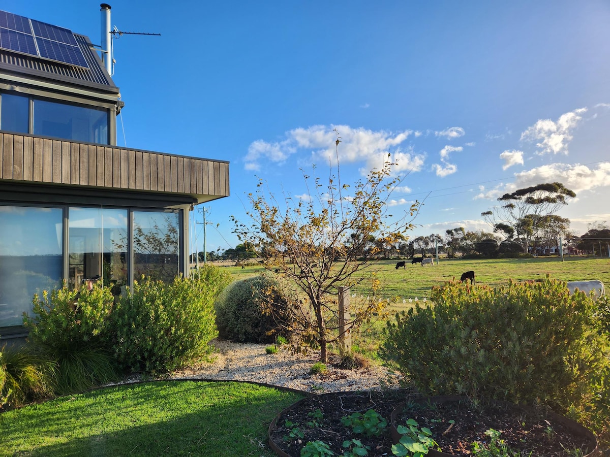 A view of the exterior garden features various plants and a small tree. Beyond the garden, farmland can be seen with grazing animals, while the sky above displays blue hues and scattered clouds. The modern home in the foreground has large windows allowing for natural light.