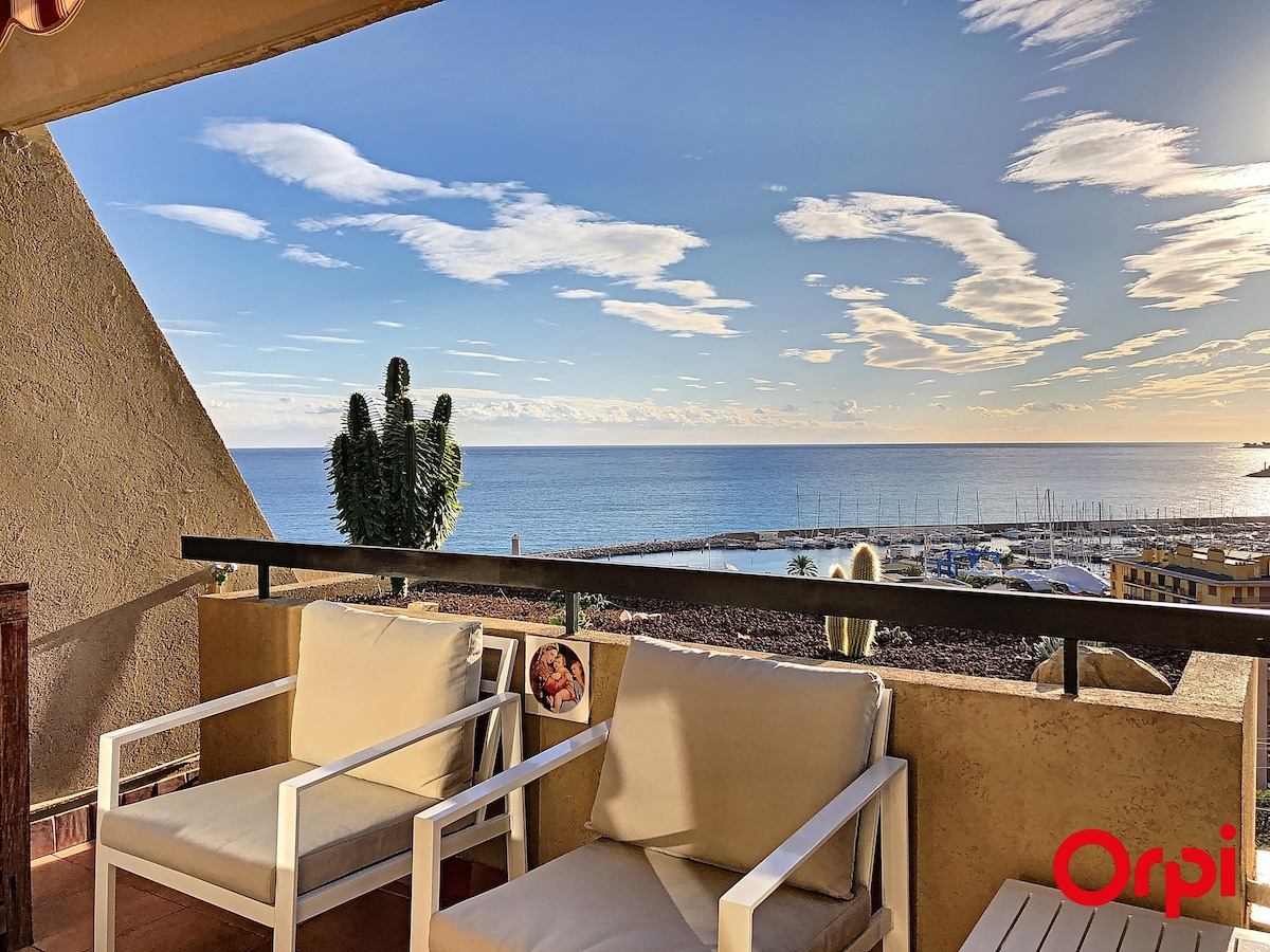 A spacious terrace is featured, showcasing two white armchairs positioned for comfort. The view overlooks the serene sea and the harbor, with the sky displaying soft clouds and bright sunlight reflecting on the water's surface.