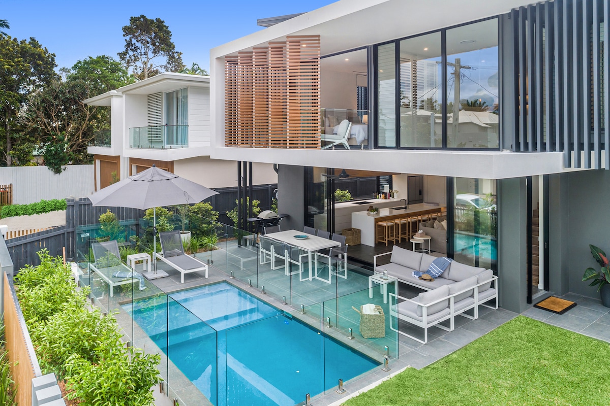 An outdoor living space features a private plunge pool, surrounded by glass fencing for safety. Comfortable seating areas are positioned nearby under an umbrella, with an outdoor dining table and chairs. The modern architecture showcases large glass windows, creating an open and inviting atmosphere.