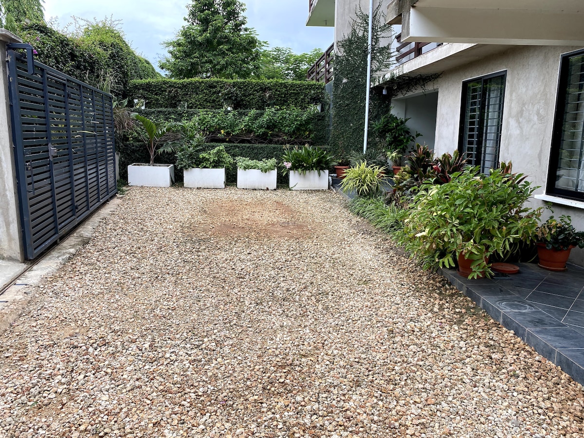 A gravel parking area is bordered by lush greenery and neatly arranged plants in decorative pots. The space is open and well-maintained, with a fence visible in the background, providing a sense of privacy and tranquility.