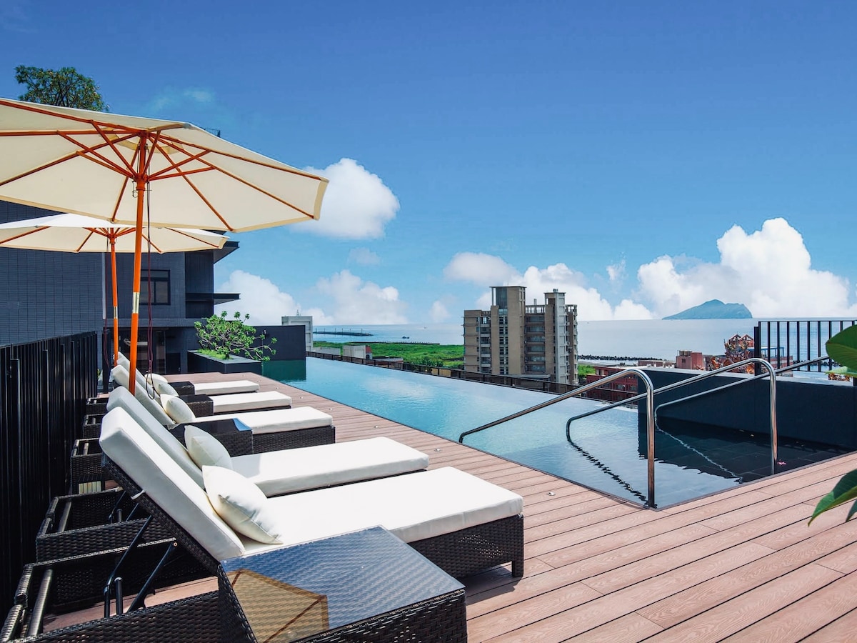 A rooftop infinity pool is surrounded by comfortable lounge chairs, each draped with soft cushions. Large umbrellas provide shade. The distant view includes mountains and a serene sea under a clear blue sky, enhancing the tranquil ambiance of the space.