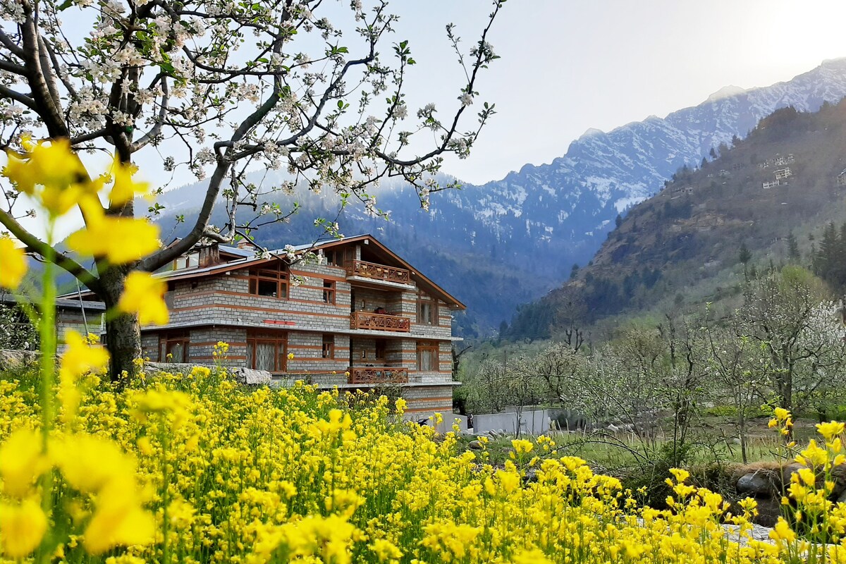 A multi-story building is situated amidst vibrant yellow flowers and surrounded by apple orchards. The backdrop features majestic mountains with snow-capped peaks. Blossoming trees add to the natural beauty of the setting, enhancing the serene environment.
