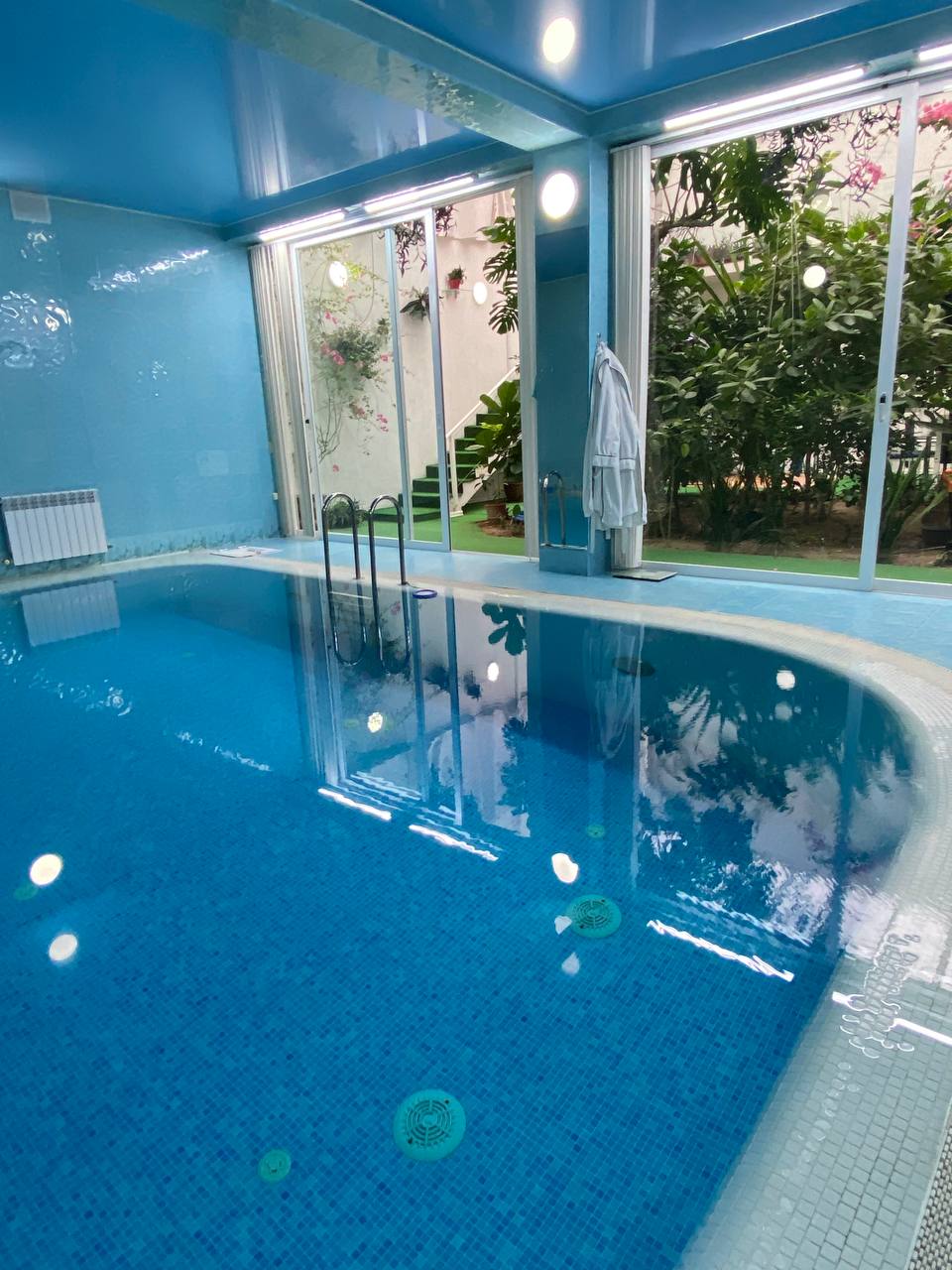 An indoor pool with clear blue water reflects soft lighting and surrounding greenery. Large glass doors invite natural light and provide views of a vibrant outdoor garden, enhancing the tranquil ambiance.