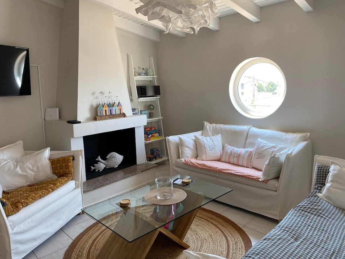 A bright living area is featured, arranged with two white sofas and a cozy circular coffee table. A unique round window allows natural light to fill the space, while a fireplace adds a functional touch. A shelf with colorful decor and small toys is also visible.