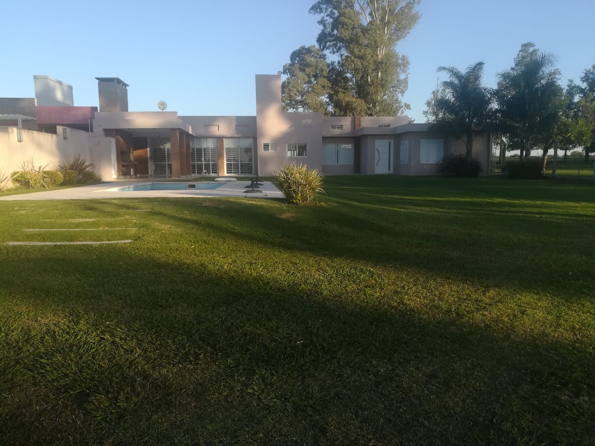 A spacious property is surrounded by lush green grass and trees. The house features large windows reflecting natural light and a classic design. A tranquil area is shown with a swimming pool, creating an inviting outdoor space.