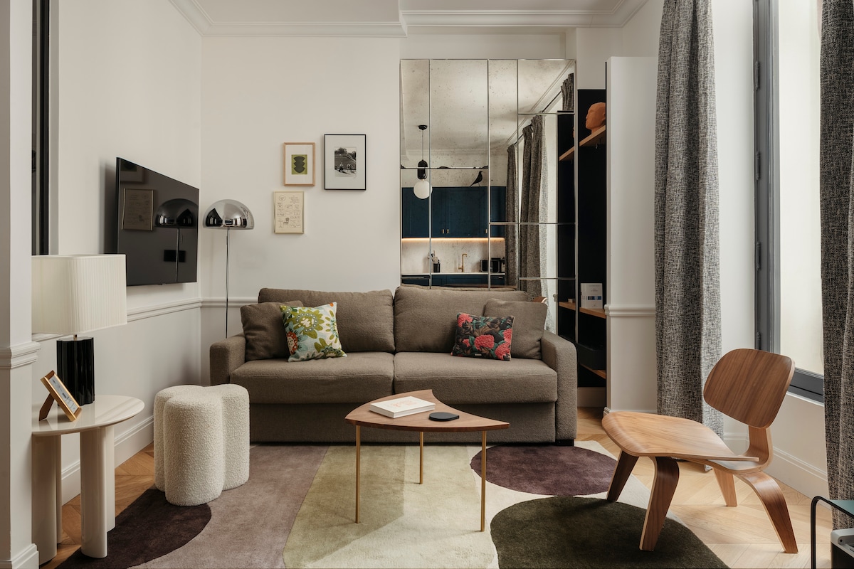 A comfortable living area is illustrated with a brown sofa adorned with decorative cushions. A modern coffee table is centered on a patterned rug. A television is mounted on the wall, with light streaming through large windows covered by flowing curtains.