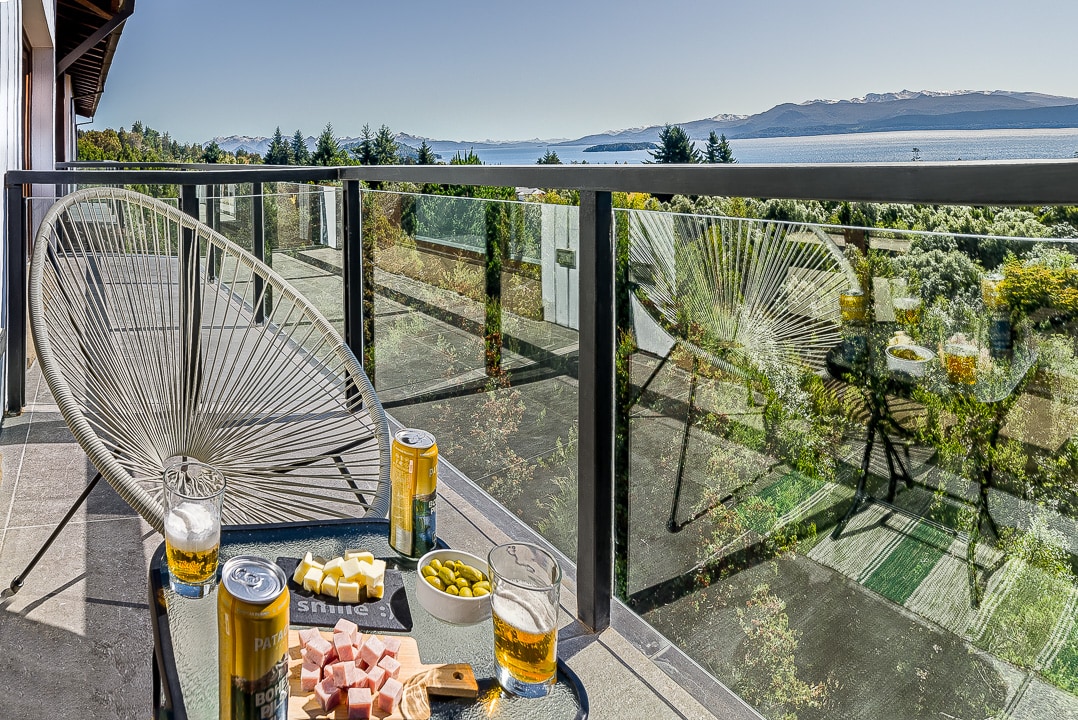 A balcony is shown with a table set, featuring a selection of cheese, olives, and drinks. Two drinks are displayed next to a bowl and a plate. The view presents distant mountains and a body of water under a clear sky, framed by glass railings.