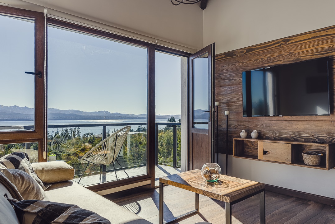 A comfortable living area is revealed, featuring large sliding glass doors that provide panoramic views of the natural surroundings. A wooden coffee table rests in front of a sofa, while a television is mounted on the wall across from the seating arrangement.