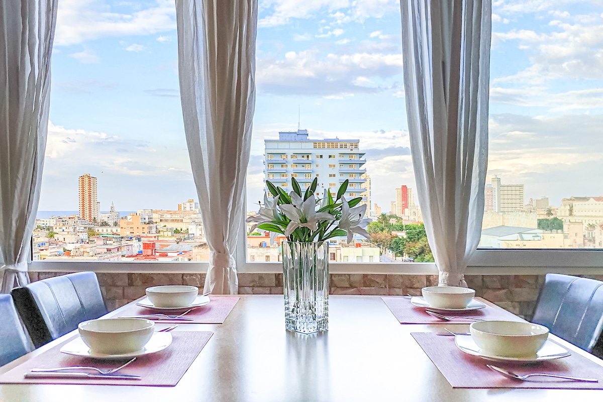 A modern dining area is presented with a sleek table set for four. Fresh white lilies are placed in a crystal-clear vase at the center, while large windows allow natural light to illuminate the space, offering city views and showcasing the vibrant skyline.