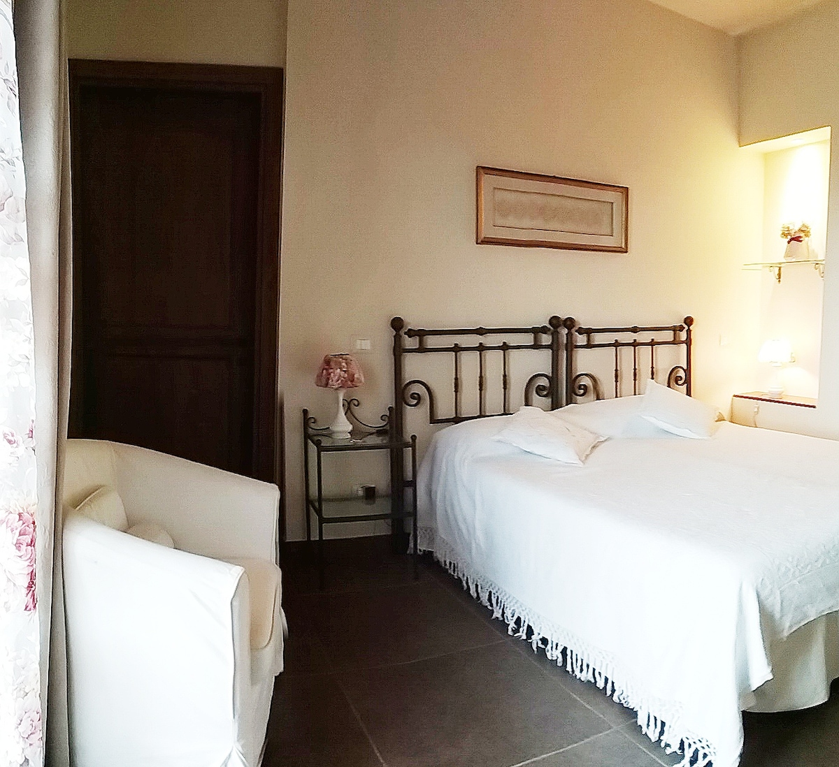 A spacious sleeping area features a classic iron bed with white linens, complemented by a soft armchair and a small table. Natural light enters through a partially open window, illuminating the neutral-toned walls and tiled flooring.