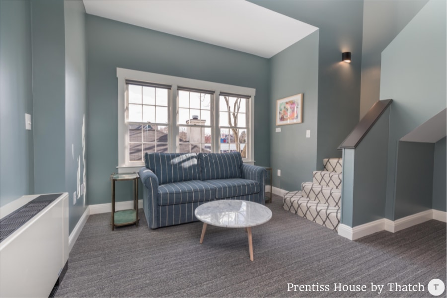 The living room features a striped sofa facing a round coffee table, with large windows providing natural light. A staircase is visible, leading to the upper level, while textured gray carpet offers a cozy touch to the space.