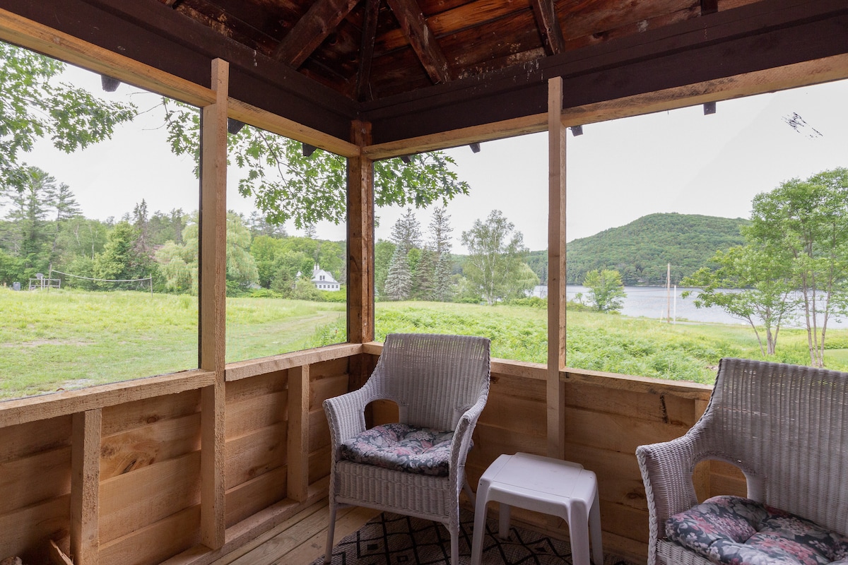 A screened porch offers views of the surrounding greenery and distant hills. Two wicker chairs with cushions are positioned alongside a small table, enhancing the inviting atmosphere of this relaxing space.