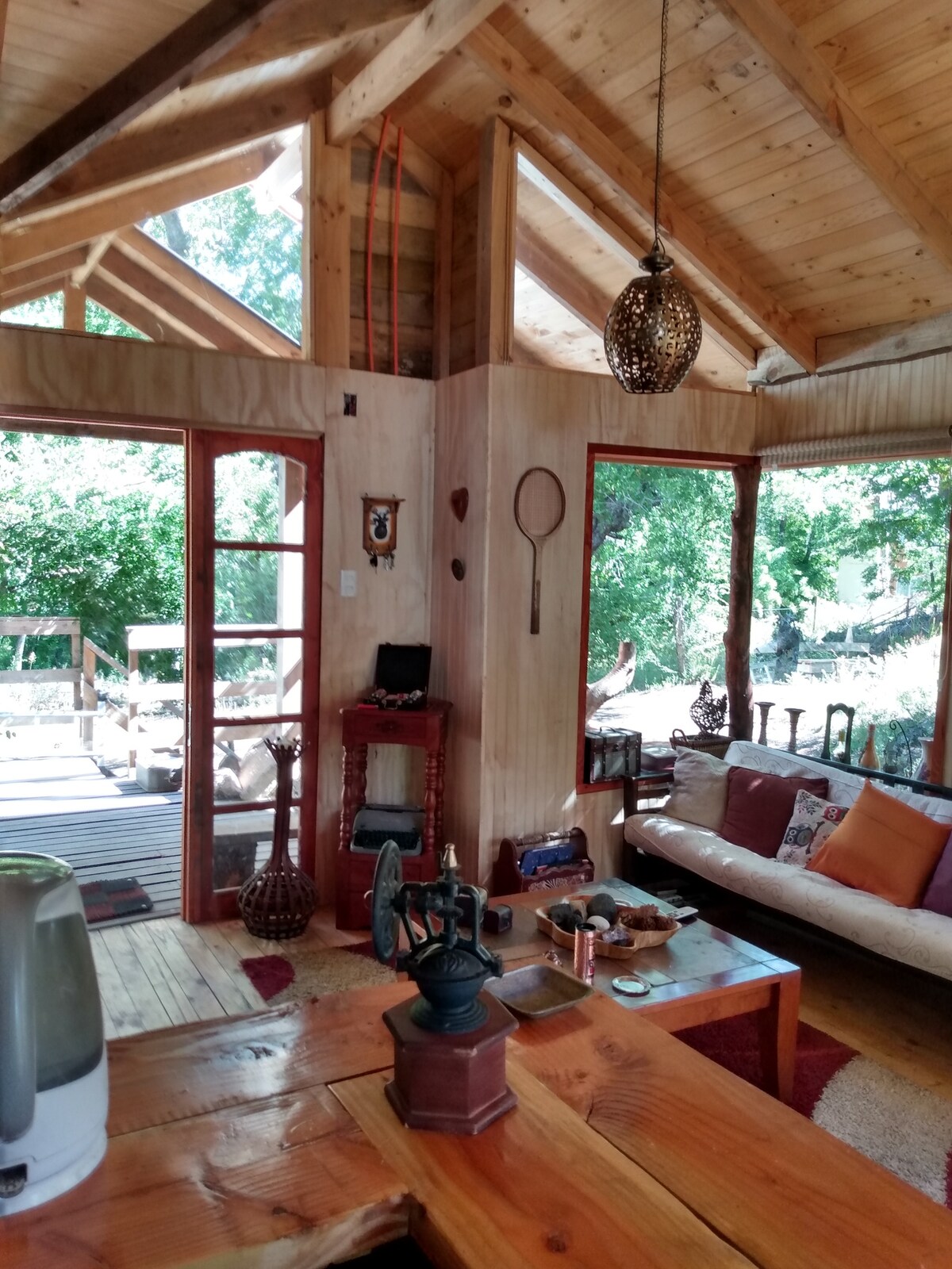 The interior of the cabin showcases natural wood finishes and high ceilings. Large windows allow ample natural light, while a sliding glass door leads to the outdoor area. Various decorative items and a cozy seating area contribute to the inviting atmosphere.