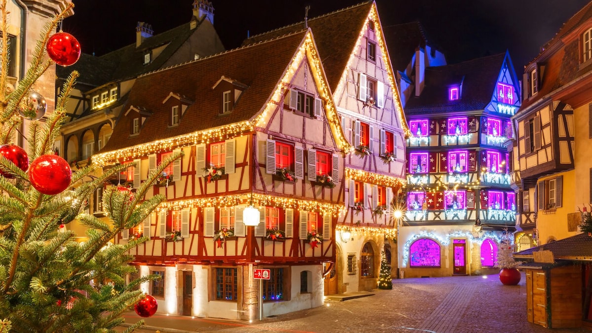 A charming street scene features traditional timber-framed buildings adorned with vibrant holiday lights. A Christmas tree decorated with red ornaments stands prominently in the foreground, while the illuminated windows of the buildings create a festive atmosphere.