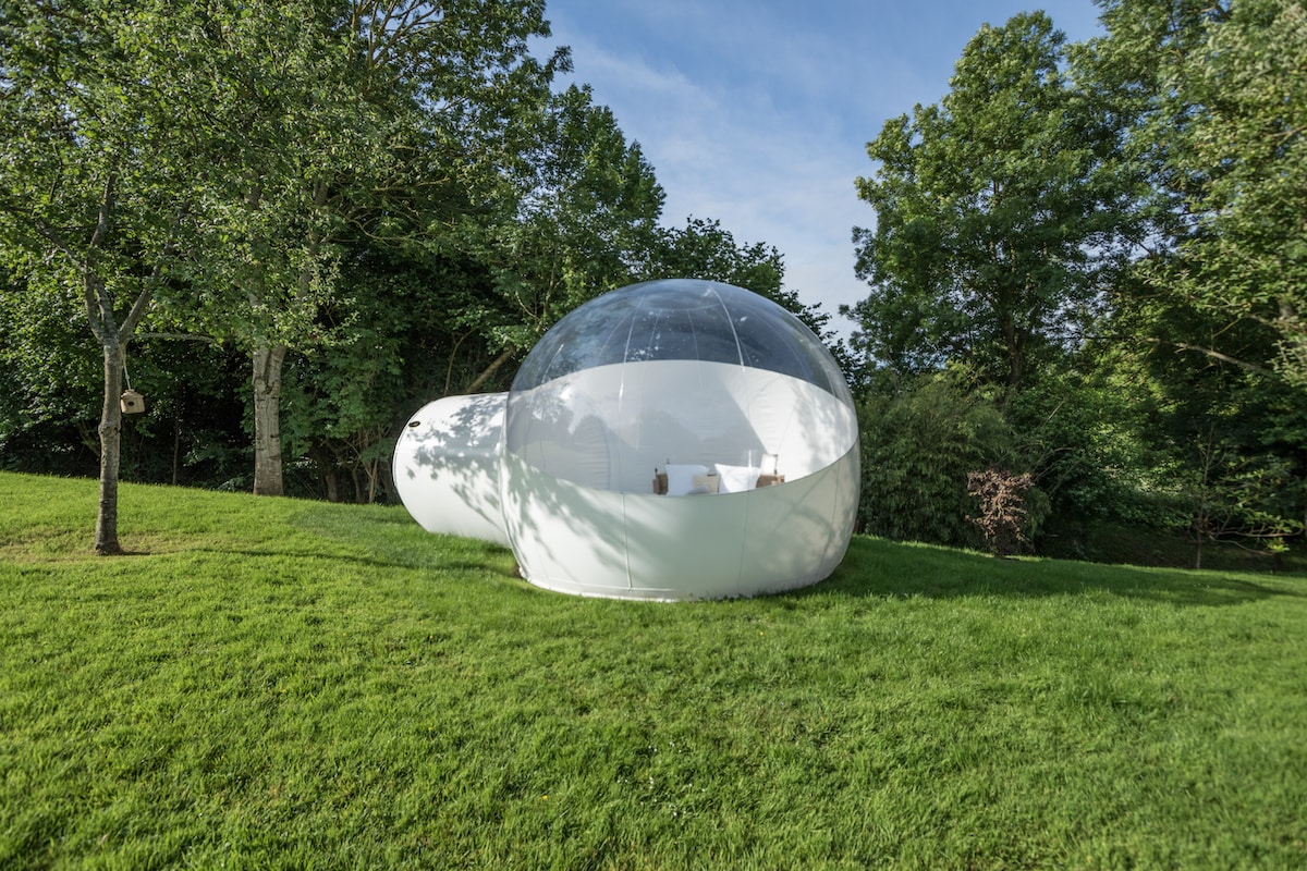 A unique transparent structure set in a lush green area, surrounded by trees. The bubble-shaped accommodation features a smooth exterior, with an undefined entrance leading inside. The layout integrates seamlessly into the natural surroundings, creating an inviting outdoor space.