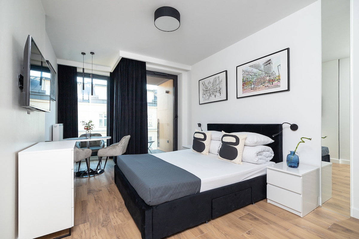 A modern studio apartment is displayed, featuring a spacious bed with a dark headboard. A small dining area with a round table and two chairs is positioned near large windows, while a TV is mounted on the wall opposite the bed. Framed artwork decorates the walls.