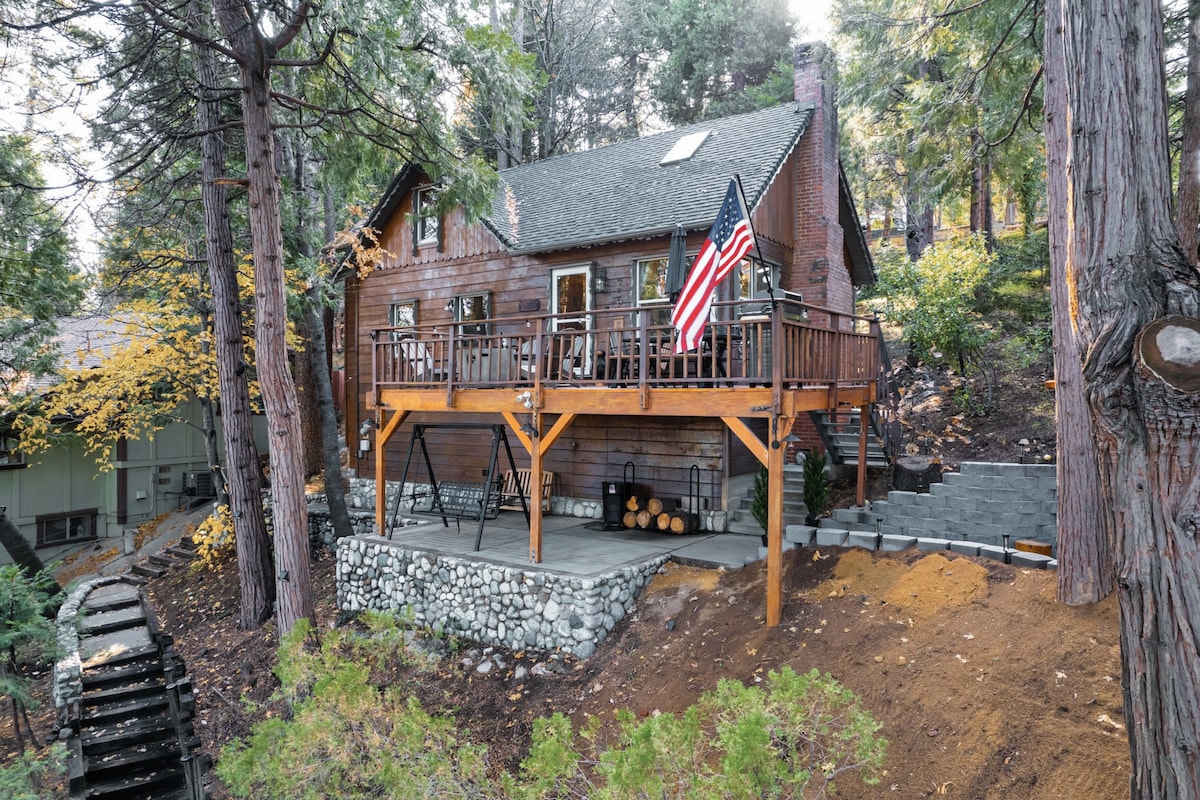 A charming two-story cabin is nestled among tall pine trees, featuring a large deck with an American flag. The exterior showcases wood siding and stone accents, with a stack of logs neatly arranged underneath. The outdoor area offers a spacious patio surrounded by natural landscaping.