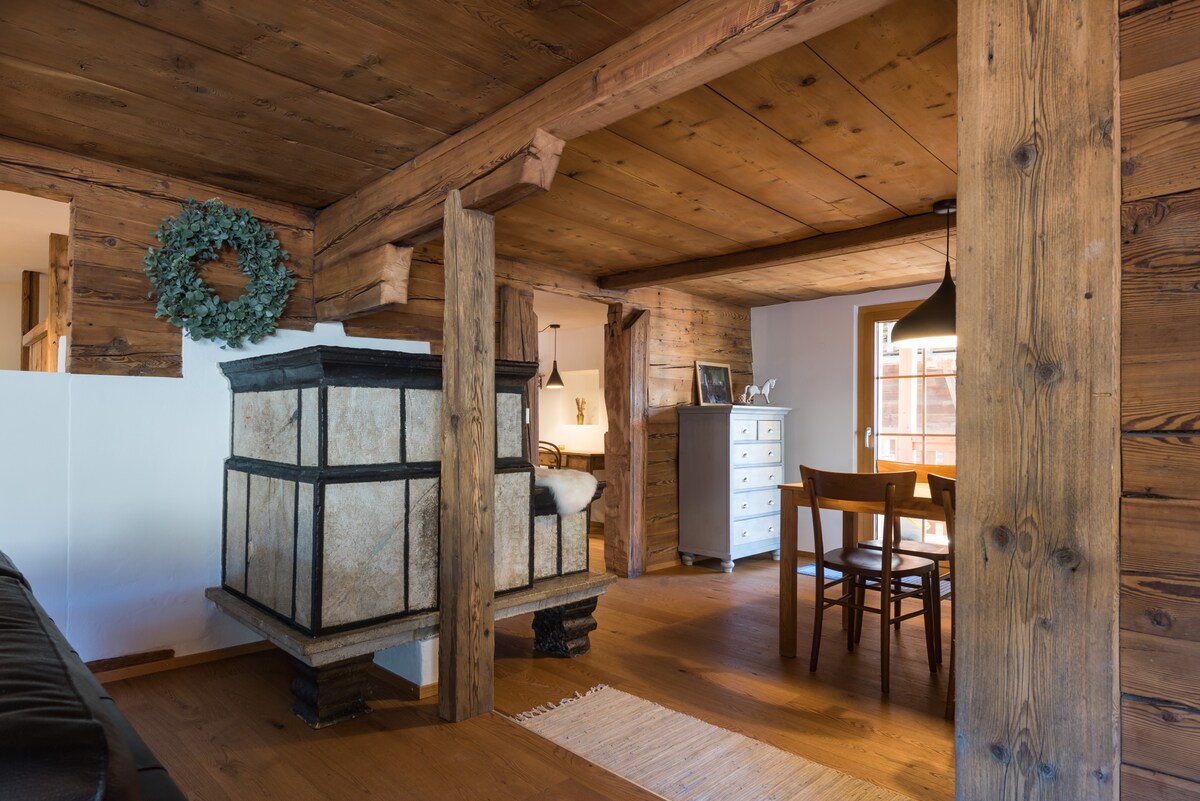 An inviting living space showcases wooden beams and a cozy fireplace adorned with stone. A dining area features a wooden table and chairs, while soft lighting creates a warm atmosphere. Natural light filters in through a window, illuminating the rustic elements of the room.