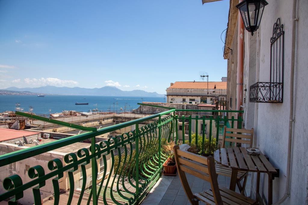 A balcony overlooking a scenic view of the sea and distant mountains includes a small wooden table and chairs. The railings are painted green, complementing the bright sky and tranquil water below, enhancing the outdoor experience of the apartment.