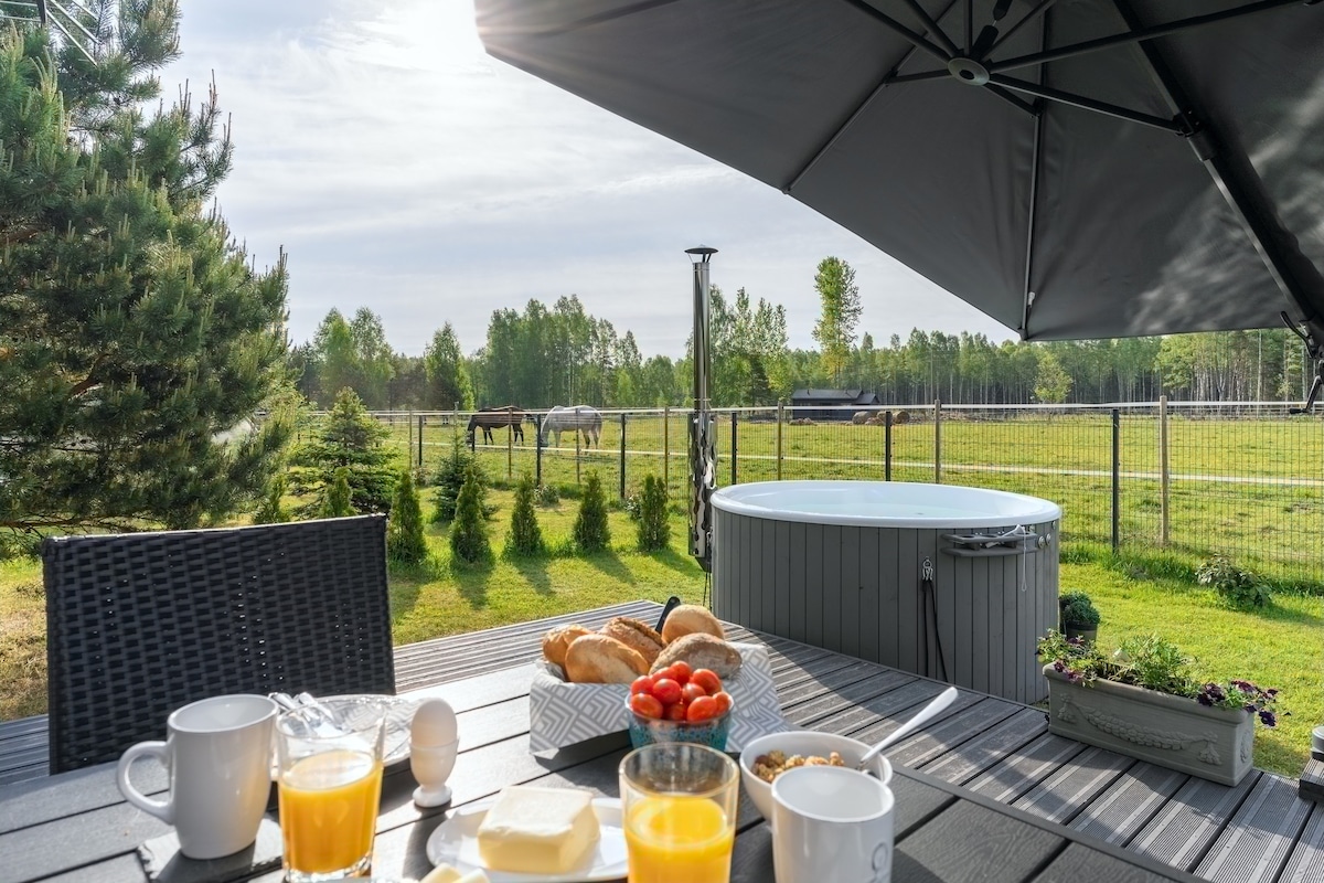 A spacious outdoor terrace is set for breakfast, featuring a table with bread, butter, fruit, and beverages. A shaded area provides comfort, while a hot tub sits nearby. In the background, a verdant field with horses grazing is visible, framed by a gentle landscape.