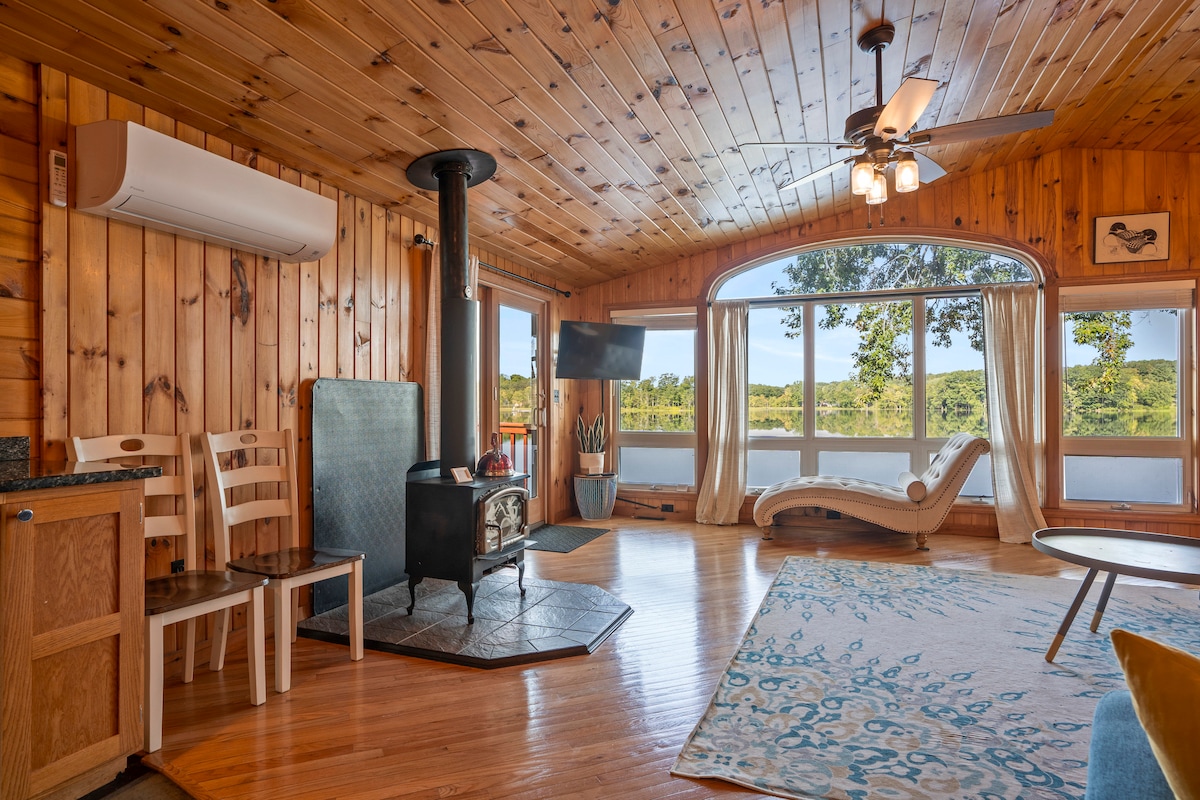 A warm wood-paneled living area features large windows offering a view of the surrounding landscape. A cozy seating area includes a lounge chair and a standing stove on a stone base. A decorative rug adds color, while a ceiling fan enhances airflow.