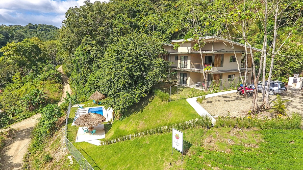 The multi-level building, set amidst lush greenery, features large balconies and open spaces. The shared pool area is visible, along with well-maintained outdoor gardens. A pathway leads from the driveway to the pool area, with surrounding trees providing natural shade.