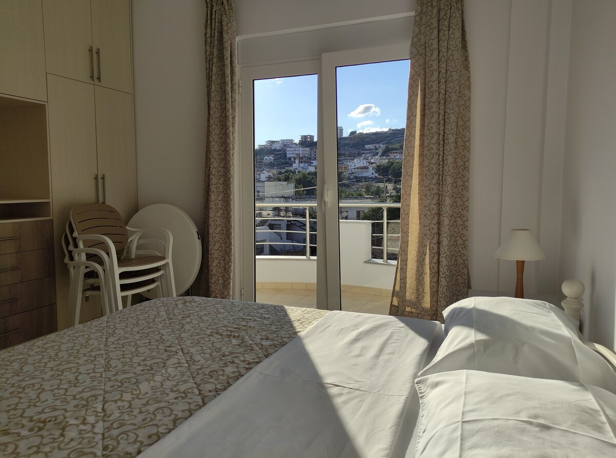 A well-lit bedroom features a spacious double bed with a light coverlet. Floor-to-ceiling windows provide access to a balcony, allowing natural light to fill the room and offering views of the surrounding landscape.
