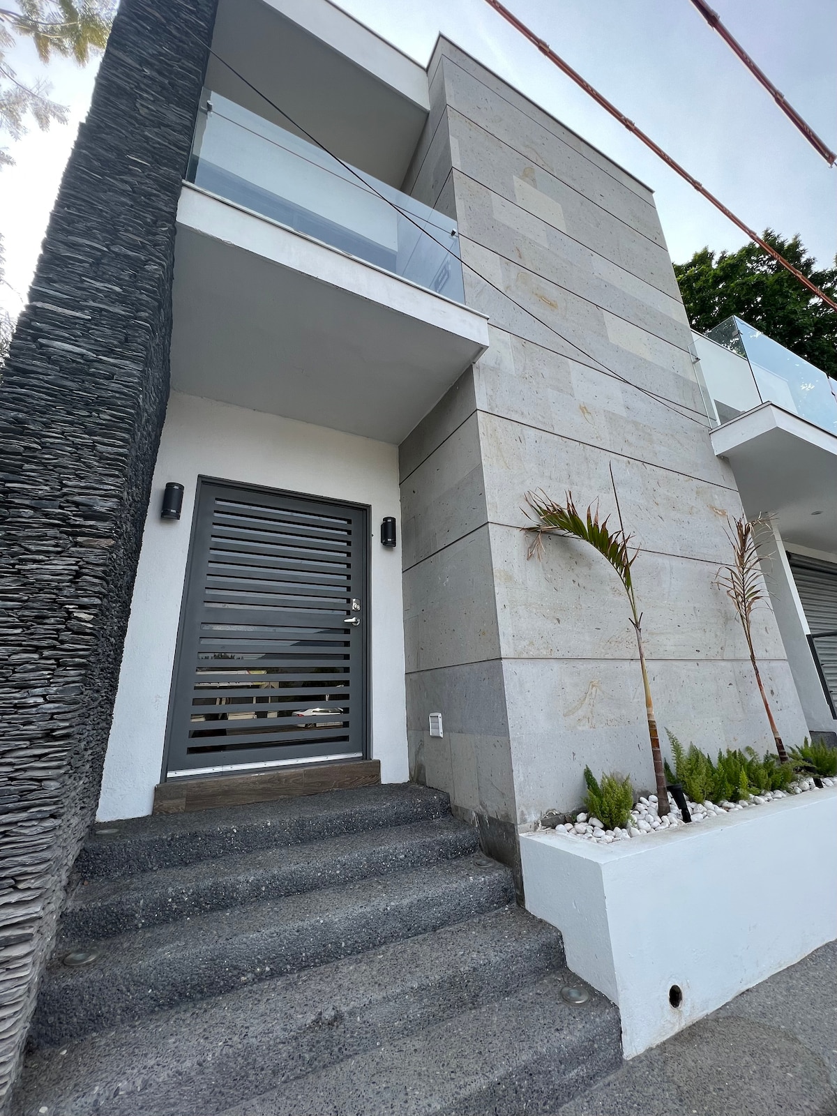 A modern facade features a sleek stone exterior with large glass balcony railings. A set of gray stone steps leads up to a minimalist door, framed by small decorative plants and white gravel along the base.