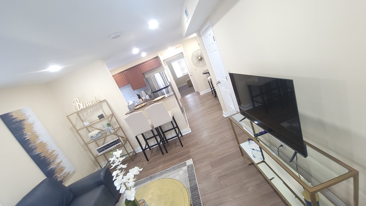 A well-arranged living space includes a dark sofa and a glass coffee table with decorative items. A modern television is positioned on a sleek console. The kitchen is visible in the background, featuring stainless steel appliances and a dining set with light-colored chairs.