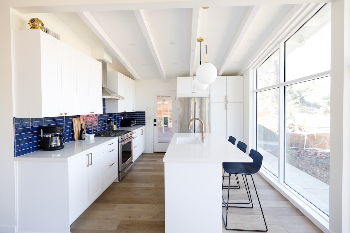 A modern kitchen features white cabinetry and a striking blue tile backsplash. A large island with seating is visible, alongside stainless steel appliances and a sleek sink. Natural light floods the space through expansive windows, creating a bright and inviting atmosphere.