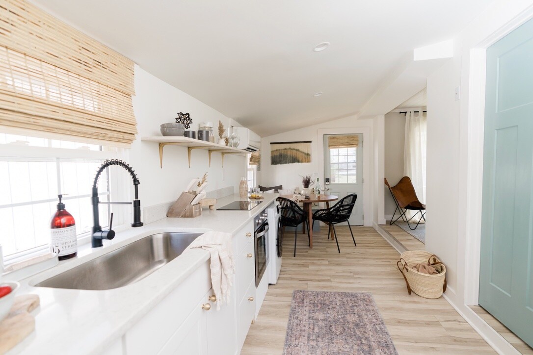 A modern kitchen features light-colored cabinetry and a spacious countertop, equipped with sleek appliances. Adjacent, a dining area includes a round table surrounded by stylish chairs. Natural light filters through window shades, illuminating the warm wood flooring.