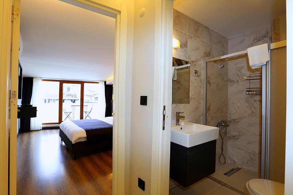 A spacious hotel room is shown through an open doorway, featuring a comfortable bed with a minimalist design. The adjacent bathroom has modern fixtures and a shower. Large windows allow natural light to fill the room, revealing a glimpse of a balcony set with two chairs.