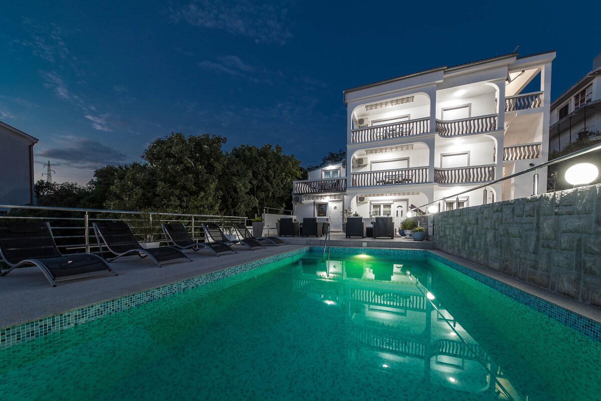 A spacious swimming pool reflects the evening sky, surrounded by lounge chairs. The building, featuring multiple balconies, is illuminated, creating an inviting atmosphere. Lush greenery is visible in the background, enhancing the tranquil setting.
