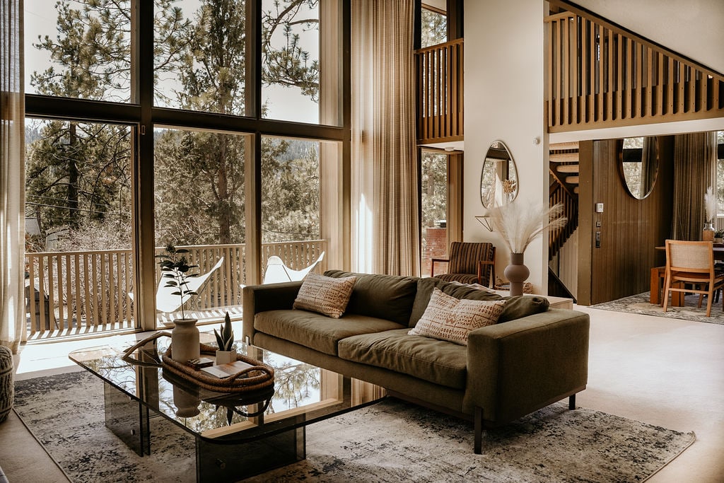A spacious living area highlights large floor-to-ceiling windows, allowing natural light to fill the room. A comfortable sofa is paired with a glass coffee table. Nearby, a dining area is visible, featuring a wooden table and chairs. The open layout creates a seamless flow to the outdoors.
