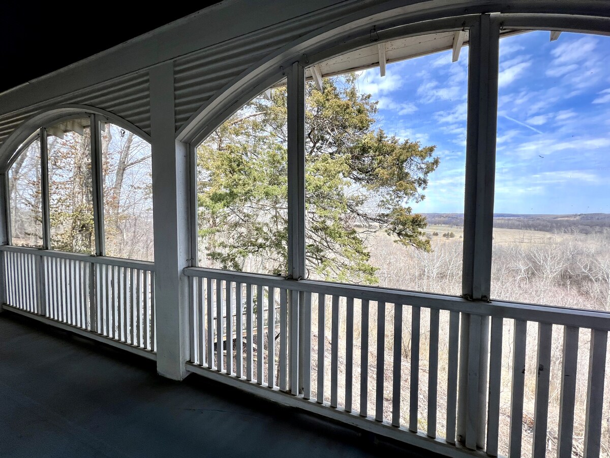 A serene view from a two-story wrap-around porch is framed by arched windows and white railings. Lush trees and rolling hills are visible beyond, with expansive blue skies adding to the tranquil atmosphere.