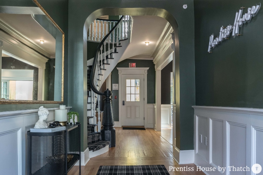 An elegant entryway features dark green walls and polished wooden flooring. A staircase, highlighted by a decorative railing, leads to the upper levels. Natural light floods in through the front door, which is framed by large windows. A mirror and minimal decor enhance the welcoming atmosphere.