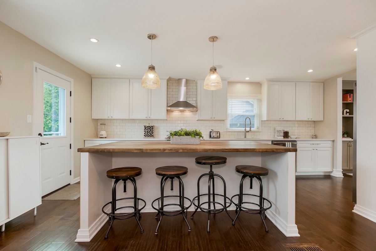 The kitchen features a large island with four round bar stools, offering ample space for gatherings. White cabinetry and light countertops complement the stainless steel appliances. Natural light floods the room through a window above the sink, enhancing the welcoming atmosphere.