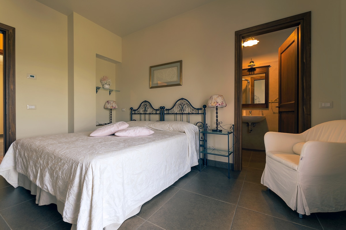 A spacious room features a large bed with a light-colored quilt, flanked by two lamps on iron nightstands. A white armchair sits in the corner, and an open doorway leads to a private bathroom, which includes stylish fixtures.