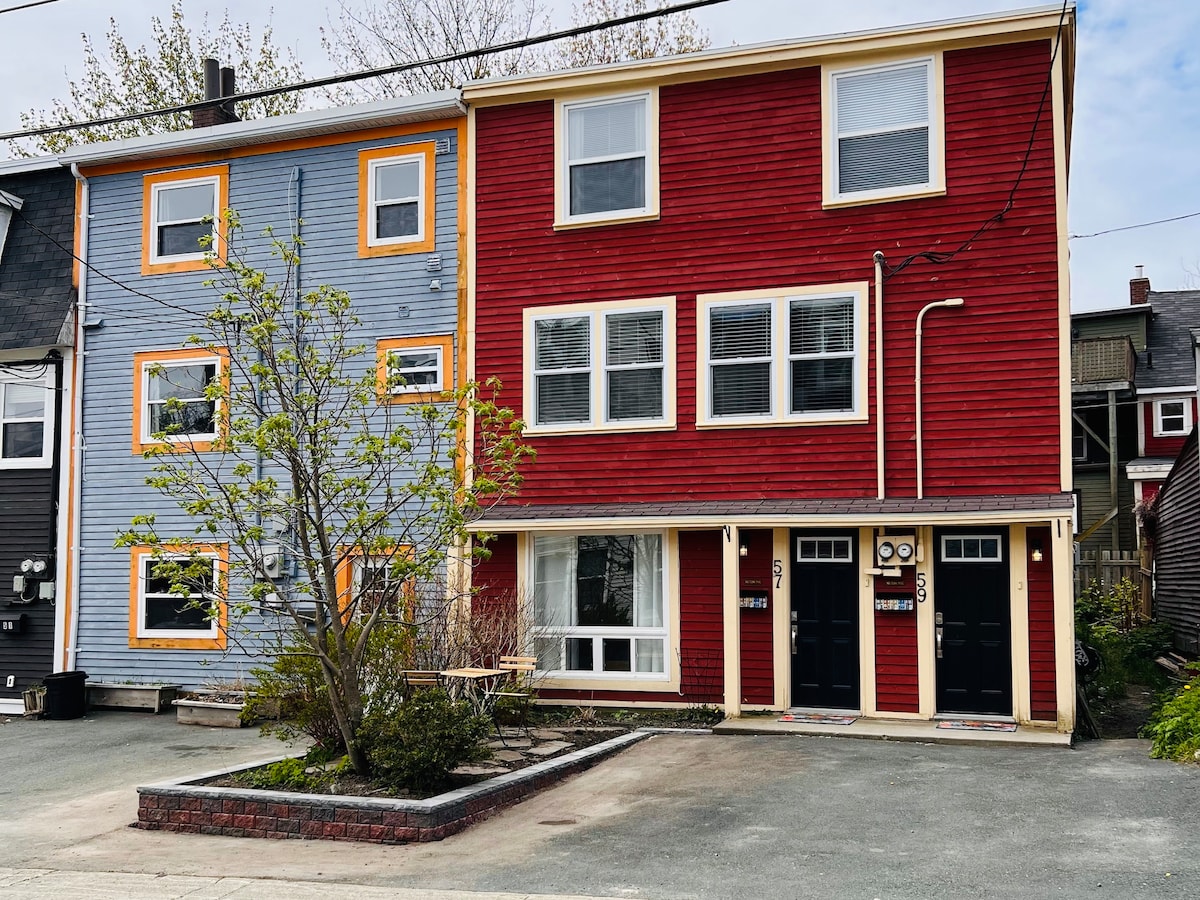 A vibrant row house features a striking red facade, complemented by cheerful blue next door. The entrance showcases two doors, framed by modest landscaping, bringing a touch of greenery to the exterior. The surrounding area is clear, providing easy access to parking.