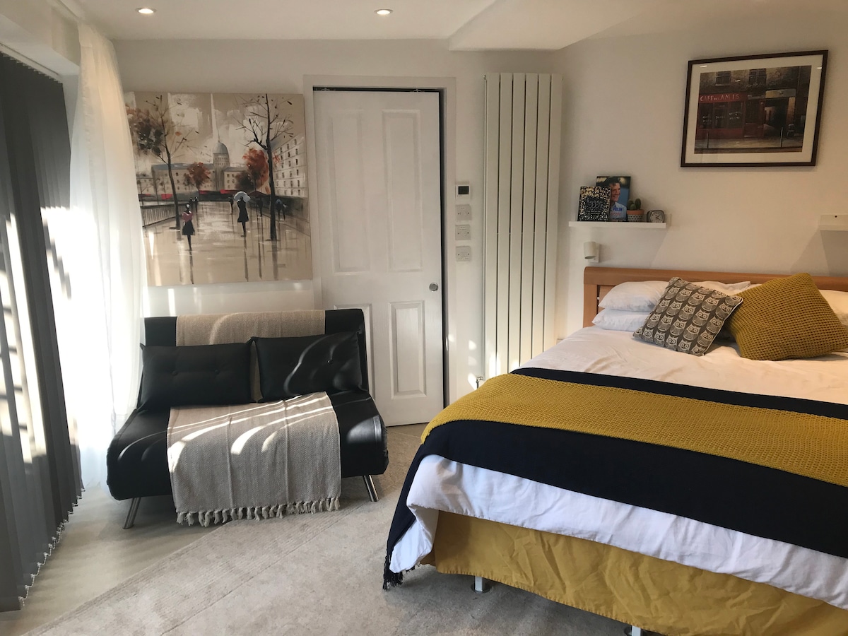 The studio room features a king size bed with navy and white bedding, complemented by a textured yellow throw. A small black sofa sits adjacent, draped with a light grey blanket. The wall is adorned with a contemporary art piece, and a door leads to an additional space.